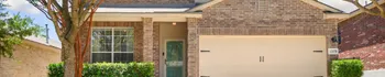 Home exterior with brick facade, attached garage, green front door, front windows, tree, and trimmed landscaping