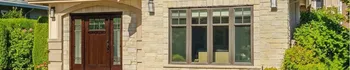 Stone home entry with wood front door, large front window, exterior wall lights, green shrubs, and detailed residential facade