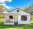 ADU builders in Los Angeles featuring a finished white backyard cottage with front porch, private entry, black shutters, lawn area, and compact accessory dwelling unit design